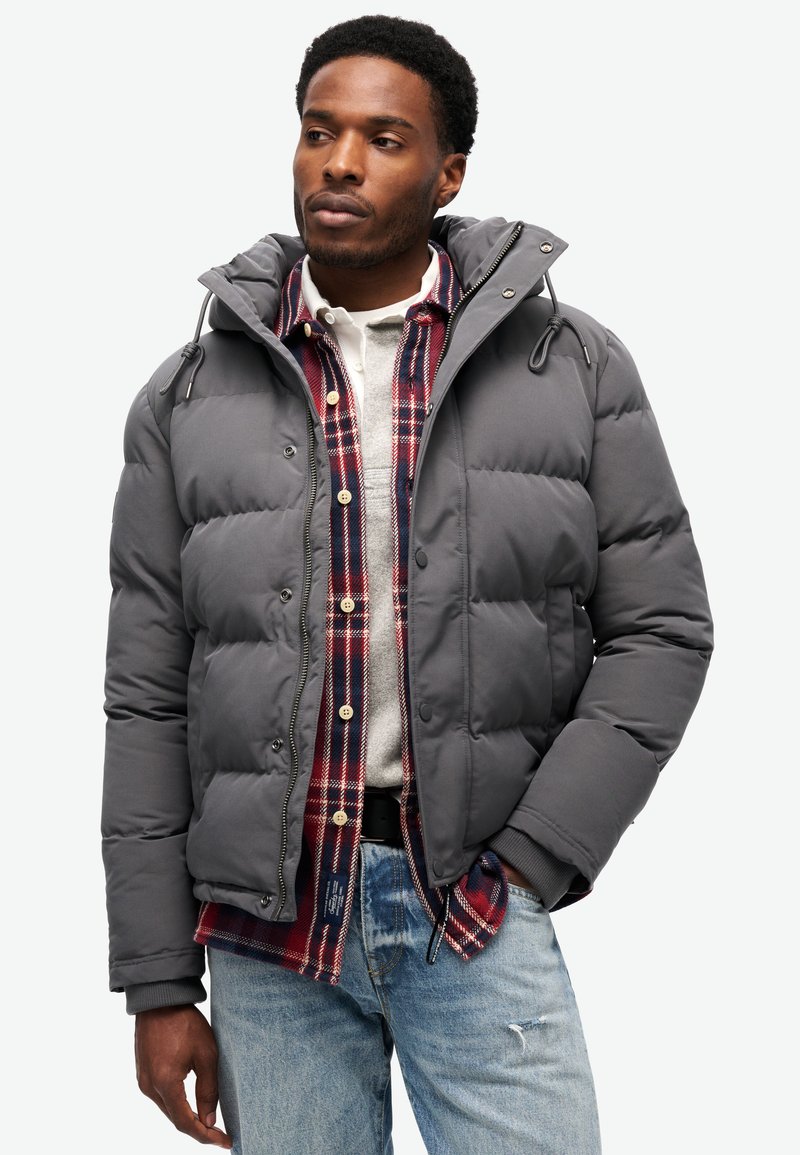Chaqueta acolchada gris con capucha, con cierre de cremallera, puños elásticos y paneles laterales en contraste, llevada sobre una camisa de cuadros y un suéter.