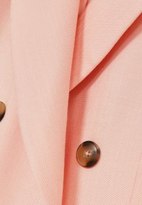 Béžový látkový blejzr s texturovanou tkaninou, zdobený klopou a dvěma hnědými knoflíky na zapínání, zdůrazňující funkční detaily.