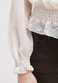 Bílá, průhledná blůza s texturovaným vzorem, elastickým pasem a volánkovými manžetami. V kombinaci s tmavě hnědými džínami.