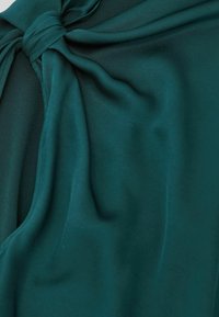 Sininerohelise satiinriie sileda tekstuuriga, millel on kaelusel sõlme detail ja pehme langevus, rõhutades voolavust ja elegantsi.