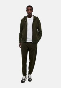 Haut à capuche vert foncé avec fermeture éclair à l'avant et poches kangourou, associé à un pantalon assorti et des baskets noir et blanc, sur un fond uni.