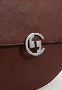 Brązowa skórzana torebka o gładkiej teksturze, z widocznym srebrnym zapięciem w kształcie logo oraz delikatnymi przeszyciami wzdłuż zamknięcia.