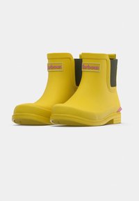 Paire de bottes de pluie jaunes courtes avec le logo "Barbour" rose sur le devant et des panneaux latéraux élastiques noirs, présentées sur fond blanc.