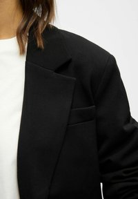 Detailní záběr na osobu v černém saku přes bílé tričko, ukazující rameno, klopu a část hnědých vlasů.