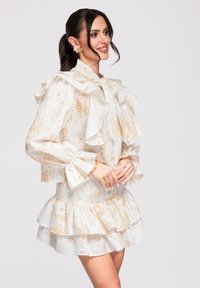 Femme portant une robe blanche à motifs floraux dorés, avec des manches à volants et une jupe à étages, souriante et posant les mains jointes.