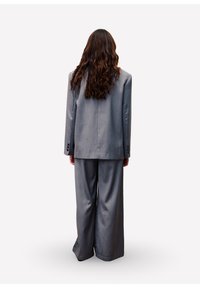 Grauer übergroßer Anzug mit langem Blazer und weit geschnittenen Hosen aus strukturiertem Stoff, von hinten vor weißem Hintergrund gezeigt.