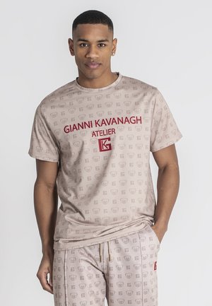 Man wearing beige Gianni Kavanagh Atelier t-shirt and matching pants with subtle logo pattern, standing with one hand in pocket.