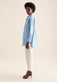 Camicia oversize azzurro chiaro con maniche lunghe, chiusura a bottoni e orlo arrotondato, abbinata a pantaloni crema a gamba affusolata e sandali neri con tacco.