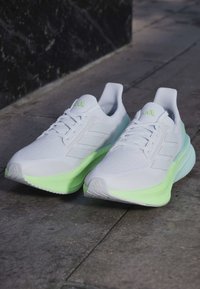 Paire de chaussures de course blanches avec semelles vert clair posées sur un pavé en pierre grise près d'un mur sombre.