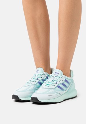 Jambes portant des baskets Adidas bleu clair avec des rayures violettes, lacets attachés, debout sur une surface blanche avec des semelles noires visibles.