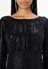 Robe noire à sequins avec manches longues, décolleté froncé et surface texturée. Présente une brillance et une silhouette ajustée.