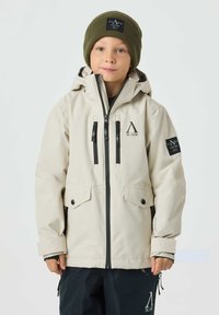 Beige wasserdichte Jacke mit Kapuze, schwarzem Reißverschluss, zwei Brusttaschen und Logodetails. Kombiniert mit einer grünen Strickmütze.