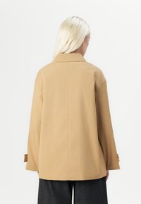 Jachetă bej, ușoară, cu o croială lejeră, guler mare și mâneci lungi. Are manșete cu nasturi și un design al cusăturii din spate drept.