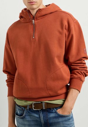 Jonge man met een roestkleurige hoodie met halve rits, een groen shirt eronder, blauwe spijkerbroek en een bruine riem met zilveren gesp.