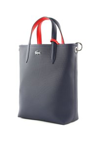 Sac fourre-tout en synthétique bleu marine avec des poignées rouges, texture lisse et accent de logo. Présente une forme structurée et un détail en métal en forme d'anneau.