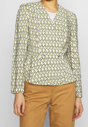 Femme portant un chemisier à manches longues à motif géométrique vert, blanc et noir avec des boutons, sur un pantalon beige, debout devant un fond uni.