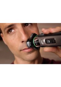 Elektrische neustrimmer in het zwart, met een groene LED-lamp en een digitaal display; de gebruiker houdt het dichtbij de neus voor precisie.