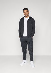 Conjunto deportivo para hombre en gris oscuro; sudadera con bolsillos frontales, camiseta blanca debajo, pantalones de chándal a juego y zapatillas blancas con detalles negros.