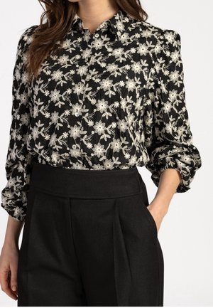 Woman wearing a black blouse with white floral embroidery and black high-waisted pants, one hand in pocket, against a white background.