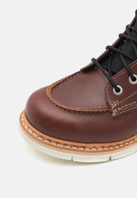 Bota de couro castanho com ponta arredondada, sola branca em contraste e atacadores pretos. Apresenta costuras visíveis e ilhós de metal.