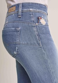 Světlé modré džíny z denimové látky s mírně opotřebovanou texturou, tradičním pětipocket designem a štítkem na pasu s názvem značky.