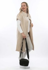 Cappotto plush beige con maniche corte, indossato sopra un top bianco e pantaloni chiari. Presenta una borsa a tracolla nera con finitura testurizzata.