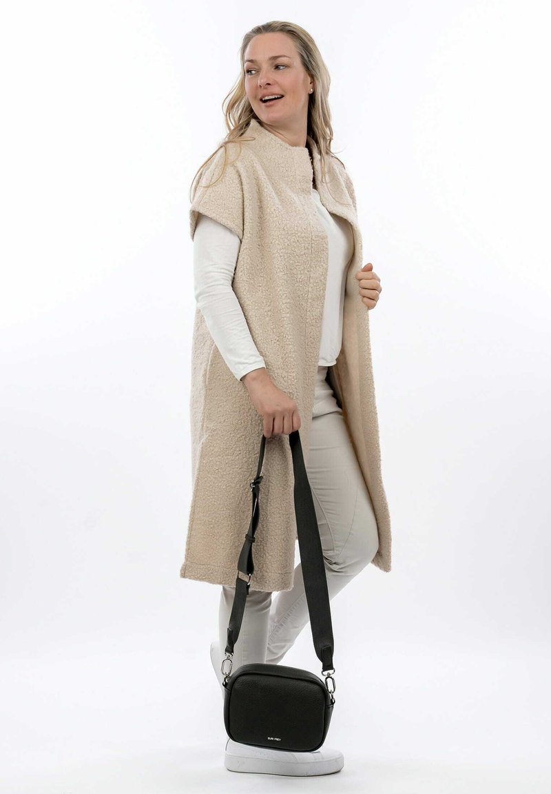 Cappotto plush beige con maniche corte, indossato sopra un top bianco e pantaloni chiari. Presenta una borsa a tracolla nera con finitura testurizzata.