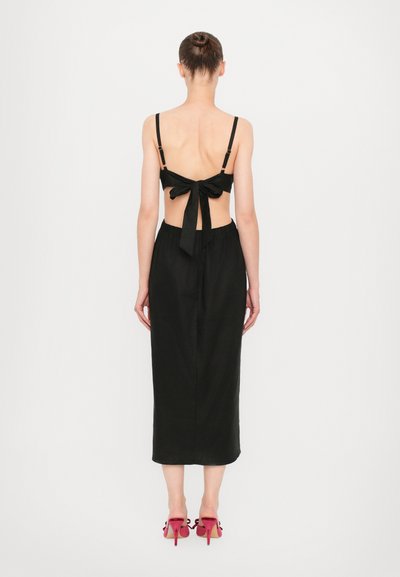 Zwarte twee-delige outfit met een rugloze top met strikdetail en een lange rok met een elastische tailleband, gecombineerd met roze hoge hakken.