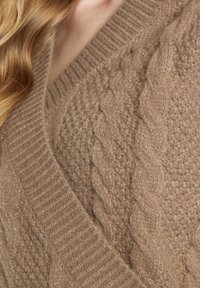 Beige gebreide trui met gestructureerde patronen, waaronder kabelbrei-details en ribbelranden, die een zachte en warme stof benadrukken.