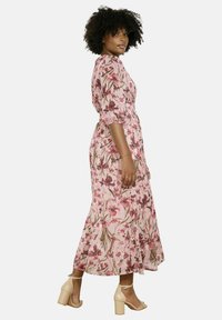 Florale maxi-jurk in roze stof met korte pofmouwen, elastische taille en een gelaagde zoom. Geaccentueerd met roze en groene bloemenpatronen.