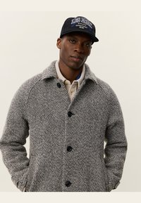 Grijze houndstooth-gepatroneerde jas met een kraag, voorknopjes en zijzakken, gedragen over een lichtgekleurde shirt en gecombineerd met een donkere pet.