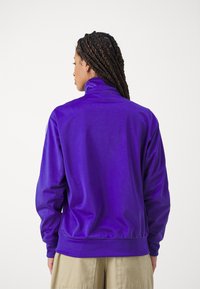 Giacca viola con zip e colletto alto, realizzata in tessuto liscio. Presenta polsini elasticizzati e una vita aderente.