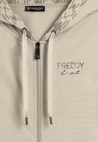 Felpa con zip di un leggero beige, con cappuccio decorato da un motivo, dotata di laccetti bianchi e dettagli in strass che formano la scritta "FREDDY dept" sul davanti.