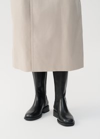 Botas de couro pretas até ao joelho, com biqueira redonda, salto bloco e fecho lateral. Usadas sob um casaco bege claro. Textura suave.