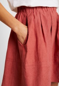 Gonna rossa al ginocchio con vita elastica e tasche laterali. Il tessuto ha una consistenza morbida e una leggera lucentezza, con pieghe e dettagli di cucitura.
