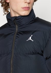 Mornarska puhovka ima visok ovratnik, zadrgo in beli Jordan logotip na prsih. Tkanina ima gladko, sijočo teksturo.