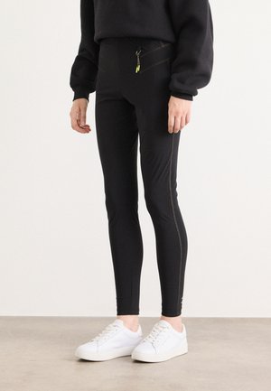 Black leggings with a smooth texture, featuring diagonal yellow stitching and a zippered pocket. Paired with white sneakers.