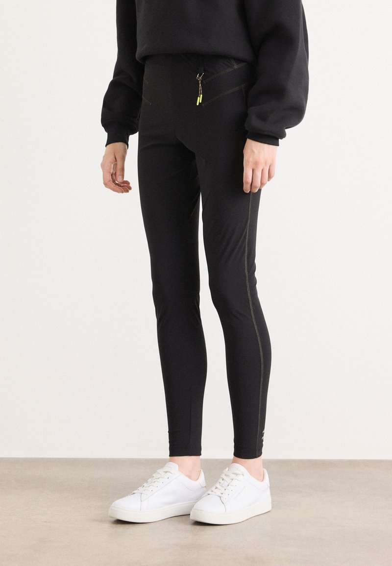 Zwarte leggings met een gladde textuur, voorzien van diagonale gele stiksels en een ritszak. Gecombineerd met witte sneakers.