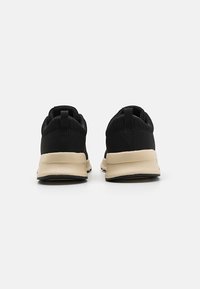 Baskets de sport noires avec une tige en maille et une semelle beige. L'arrière présente une languette de tirage pour un enfilage facile. Semelle en caoutchouc pour une meilleure traction.