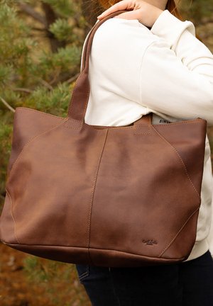 Brown leather tote bag with smooth texture, stitched accents, and wide straps, held against a light-colored sweatshirt.
