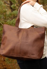 Hnědá kožená taška přes rameno s texturovaným povrchem, charakterizovaná silným šitím, prostorným designem a pevným madlem.