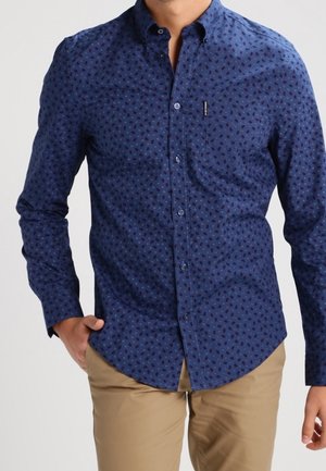 Uomo che indossa una camicia blu navy aderente con piccoli motivi scuri e pantaloni beige, mano in tasca, su uno sfondo semplice.