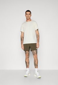 Light beige athletic t-shirt with short sleeves, paired with olive green shorts. The outfit features white sneakers and visible tattoos on the legs.