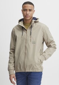 Beige zip-up jakke med hette, med en glidelås brystlomme, snor, og elastiske mansjetter. Bært over en grønn skjorte, med blå jeans.