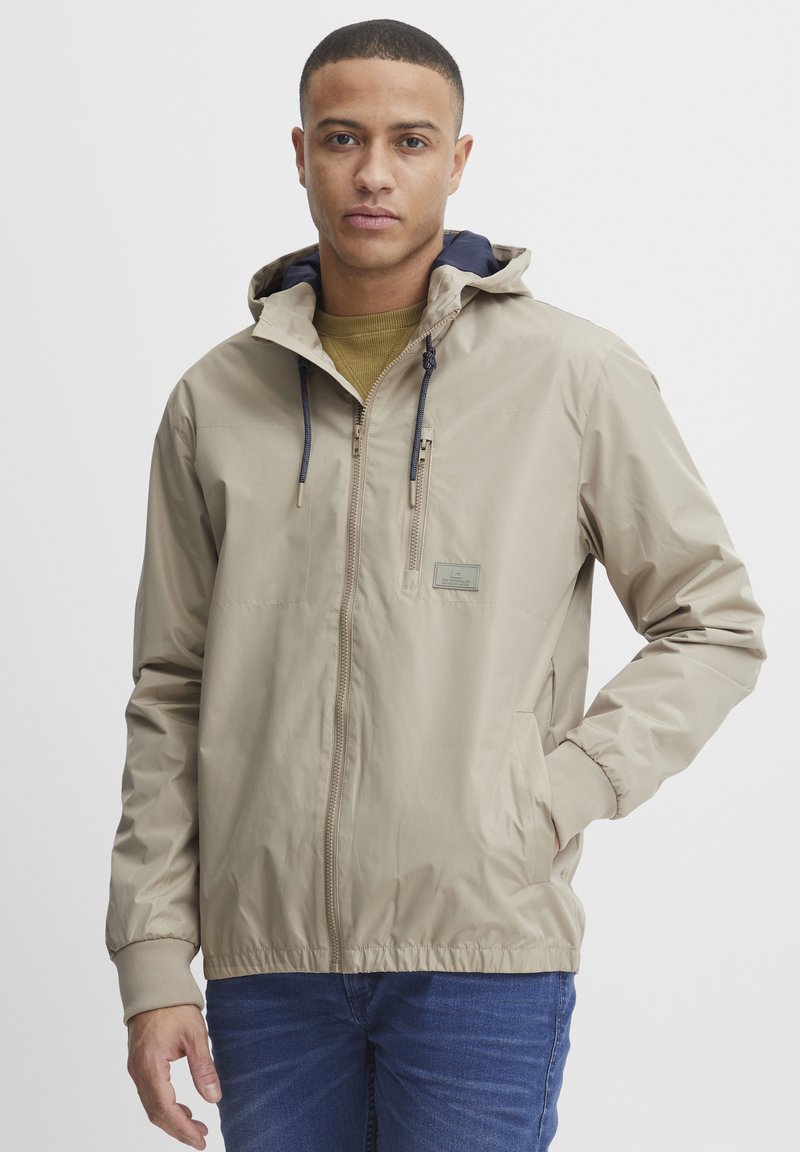 Beige zip-up jakke med hette, med en glidelås brystlomme, snor, og elastiske mansjetter. Bært over en grønn skjorte, med blå jeans.