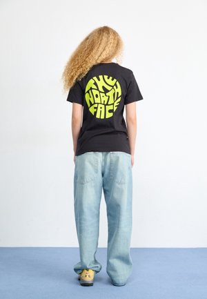 Osoba z długimi kręconymi włosami ubrana w czarną koszulkę z żółtym logo "The North Face", luźne jasnoniebieskie dżinsy i żółte trampki.