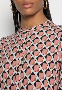Košile s geometrickým vzorem a vysokým límcem, obsahující růžové, černé a bílé barvy. Přední knoflíky a hladká, lehká látka.