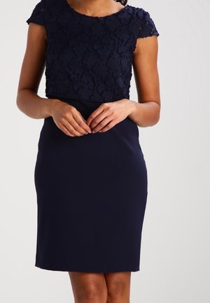 Kobieta w granatowej sukience do kolan z teksturowaną kwiatową górą i dopasowaną spódnicą, stojąca z delikatnie splecionymi rękami.