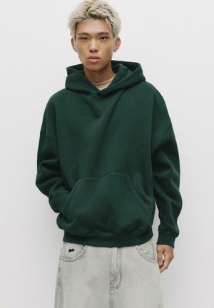 Haut de survêtement vert surdimensionné avec une poche kangourou et une capuche à cordon, fabriqué dans un matériau doux. Associé à un short en denim gris clair.