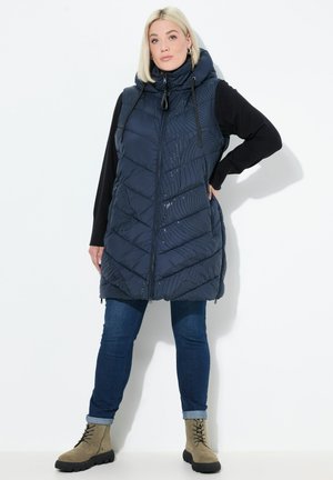 Donna che indossa un gilet imbottito senza maniche di colore blu navy con cappuccio, maniche lunghe nere, jeans blu e stivali beige con lacci, in piedi contro uno sfondo bianco.